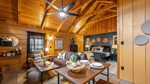 A cozy log cabin interior with a dining area, a living room with a sofa and armchairs, a kitchen area, and a bedroom with a bed and nightstands. - Location-quiet-hottub-fireplace (Sevierville)