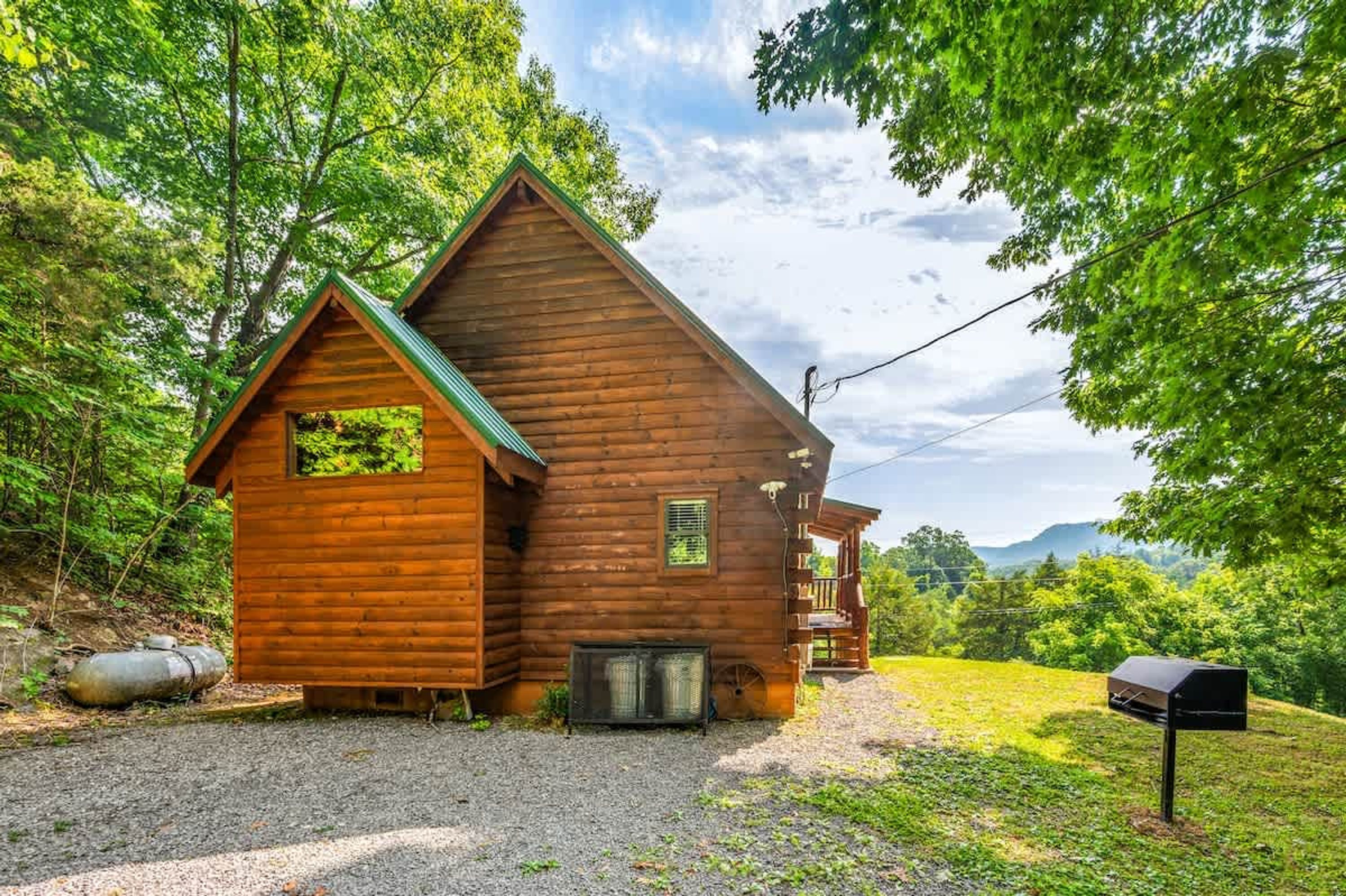 A wooden cabin with a porch, surrounded by trees and a gravel driveway.