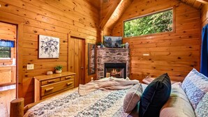 Un acogedor dormitorio de cabaña con chimenea de piedra, paredes de madera, una cama con colcha floral y una ventana con vistas a la vegetación.