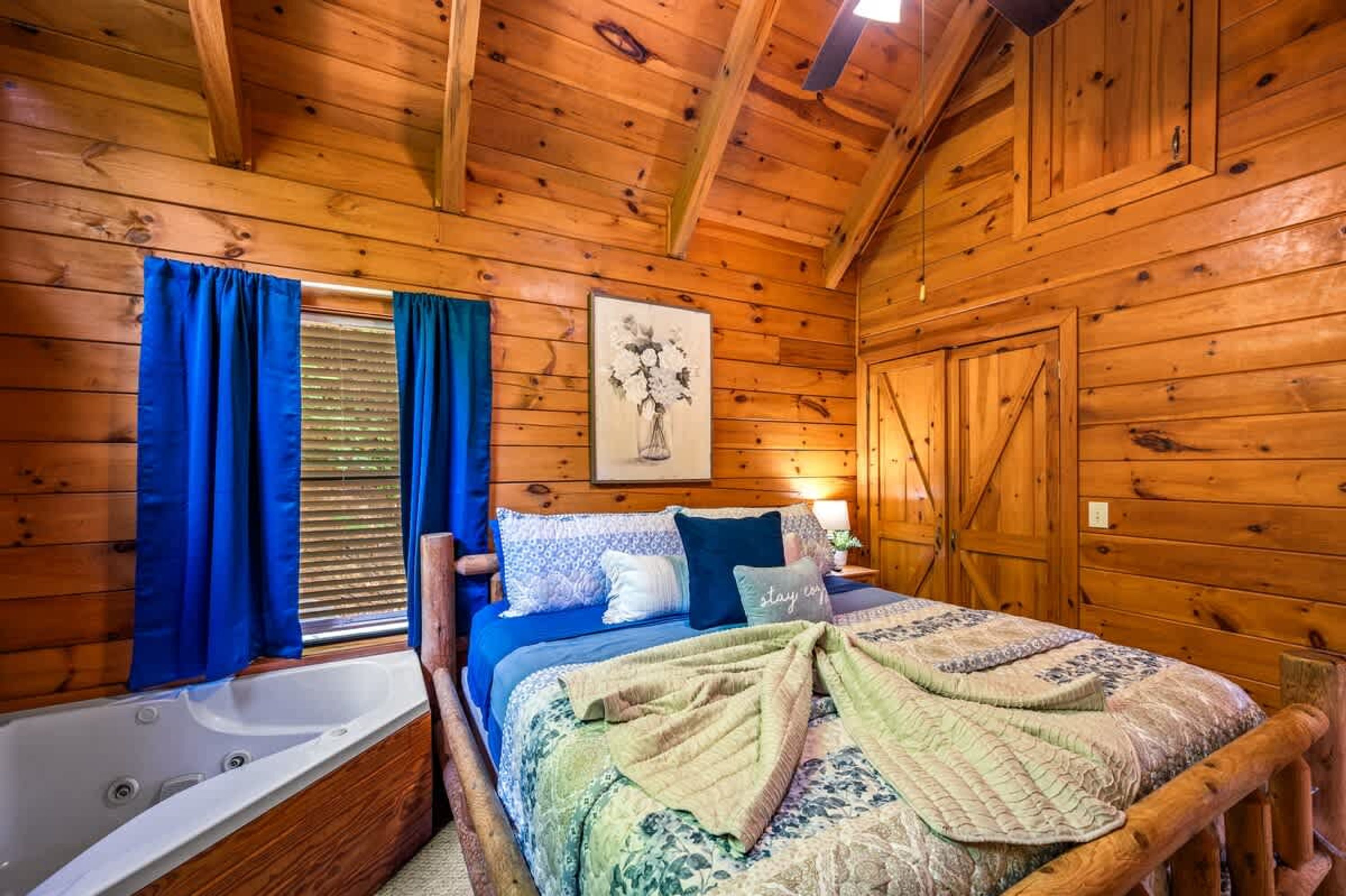 A wooden cabin bedroom with a bed, a window with blue curtains, a painting on the wall, and a wooden door.