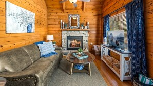 Interior acogedor de una cabaña de troncos con chimenea de piedra, ventilador de techo y televisor sobre un soporte de madera.
