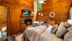 A cozy cabin interior with a wooden interior, a fireplace, a bed with pillows, a television, and a ceiling fan. - View-location-hot Tub-pool Table-3 Fireplaces (Sevierville)
