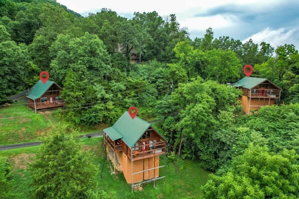 Three wooden cabins with green roofs, surrounded by dense forest, each with a red location marker. - View-location-hot Tub-pool Table-3 Fireplaces (Sevierville)
