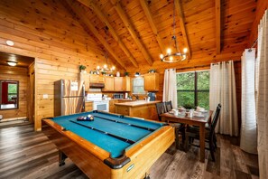 Interior de una cabaña de madera con mesa de billar, cocina, comedor y ventanas amplias.