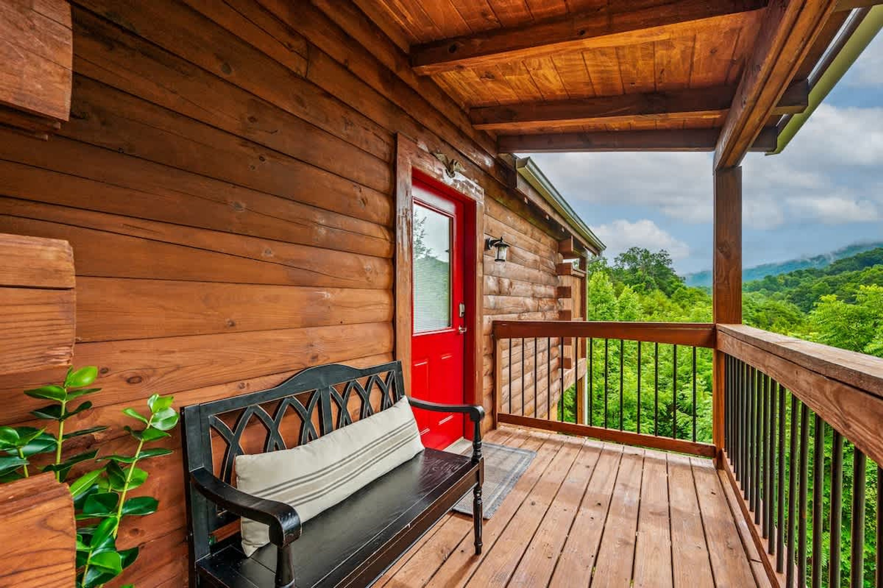 Una cabaña de madera con puerta roja, una banca y un balcón con vistas a un paisaje boscoso.