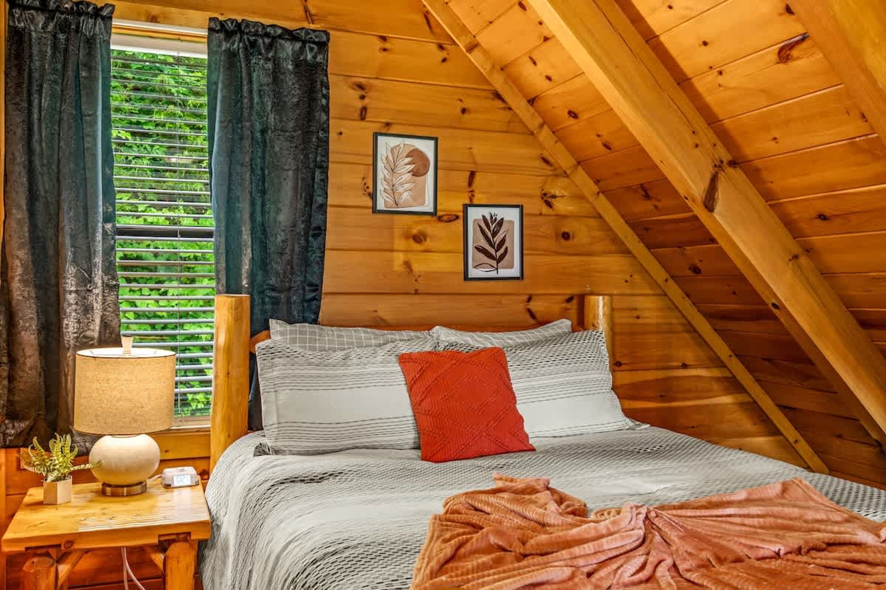 A cozy wooden room with a bed, a nightstand with a lamp, and framed botanical prints on the wall.