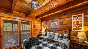 Un dormitorio de cabaña de madera con una cama grande, una mesita de noche, una lámpara y una ventana con persianas.