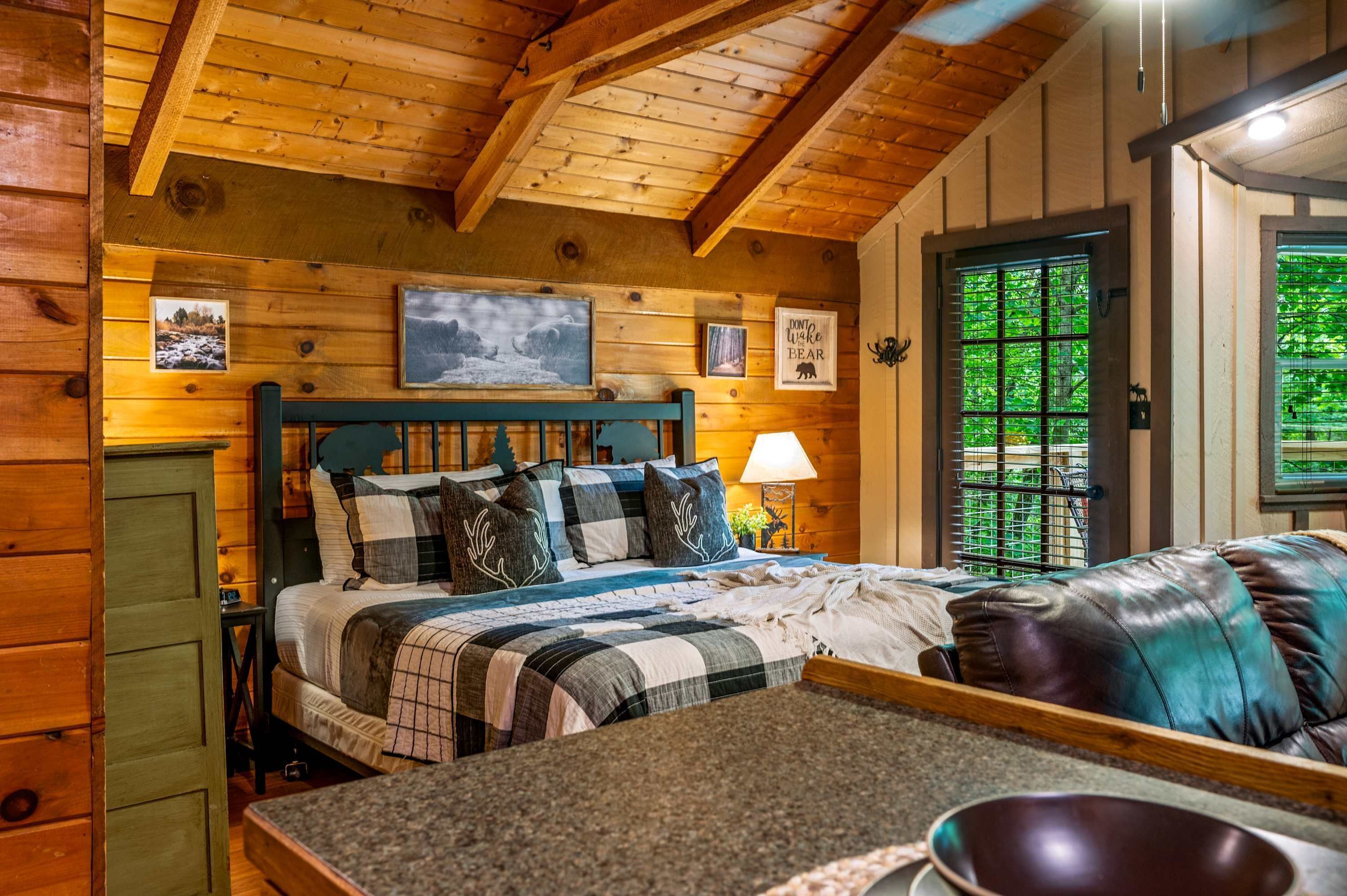 A wooden cabin interior with a bed, a dining table, and a view of greenery outside.