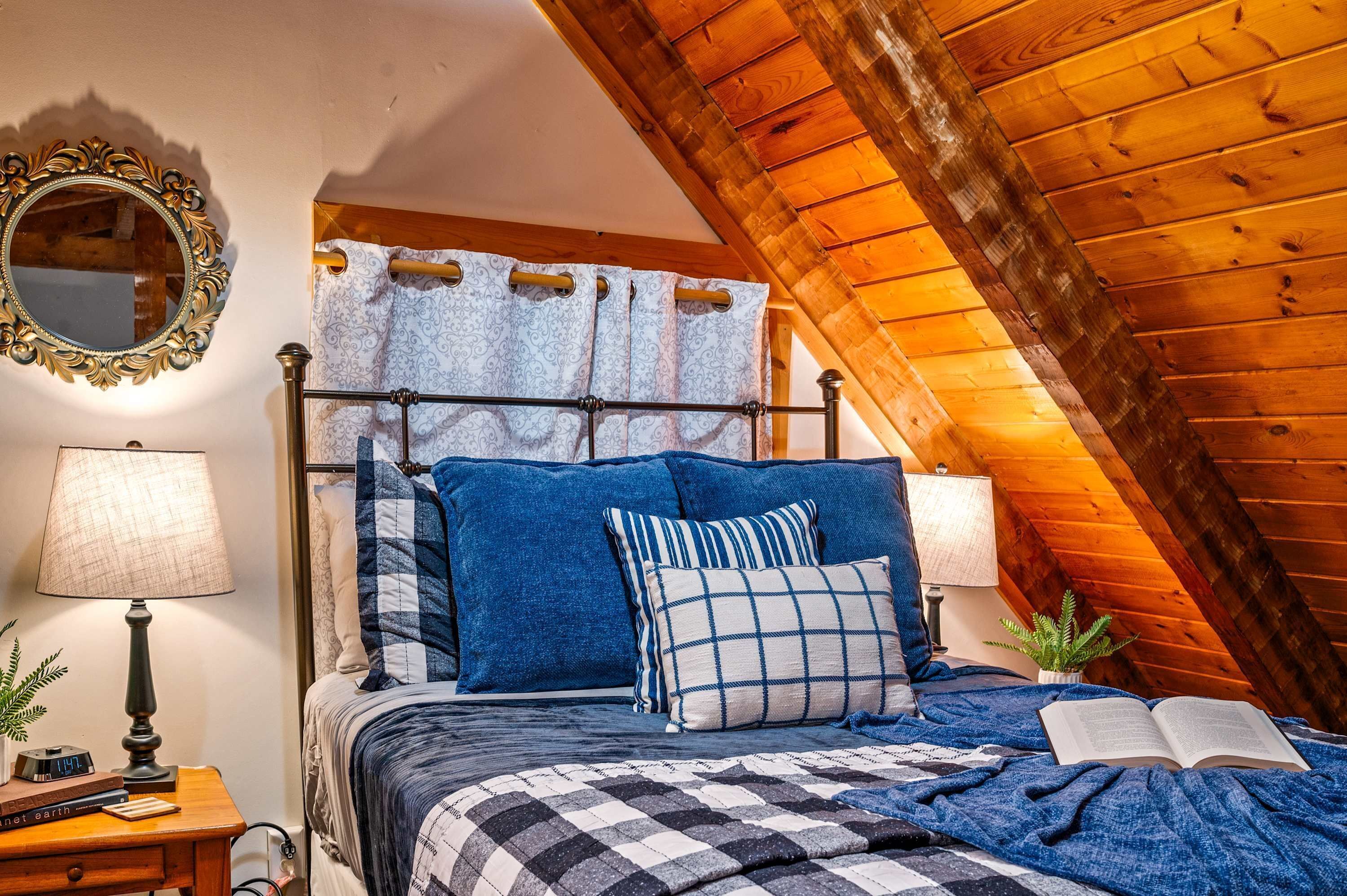 Ein Schlafzimmer mit schrägem Dach, einem Bett mit blauer Bettwäsche, einer Nachttischlampe, einem Spiegel und einer kleinen Pflanze auf dem Nachttisch.