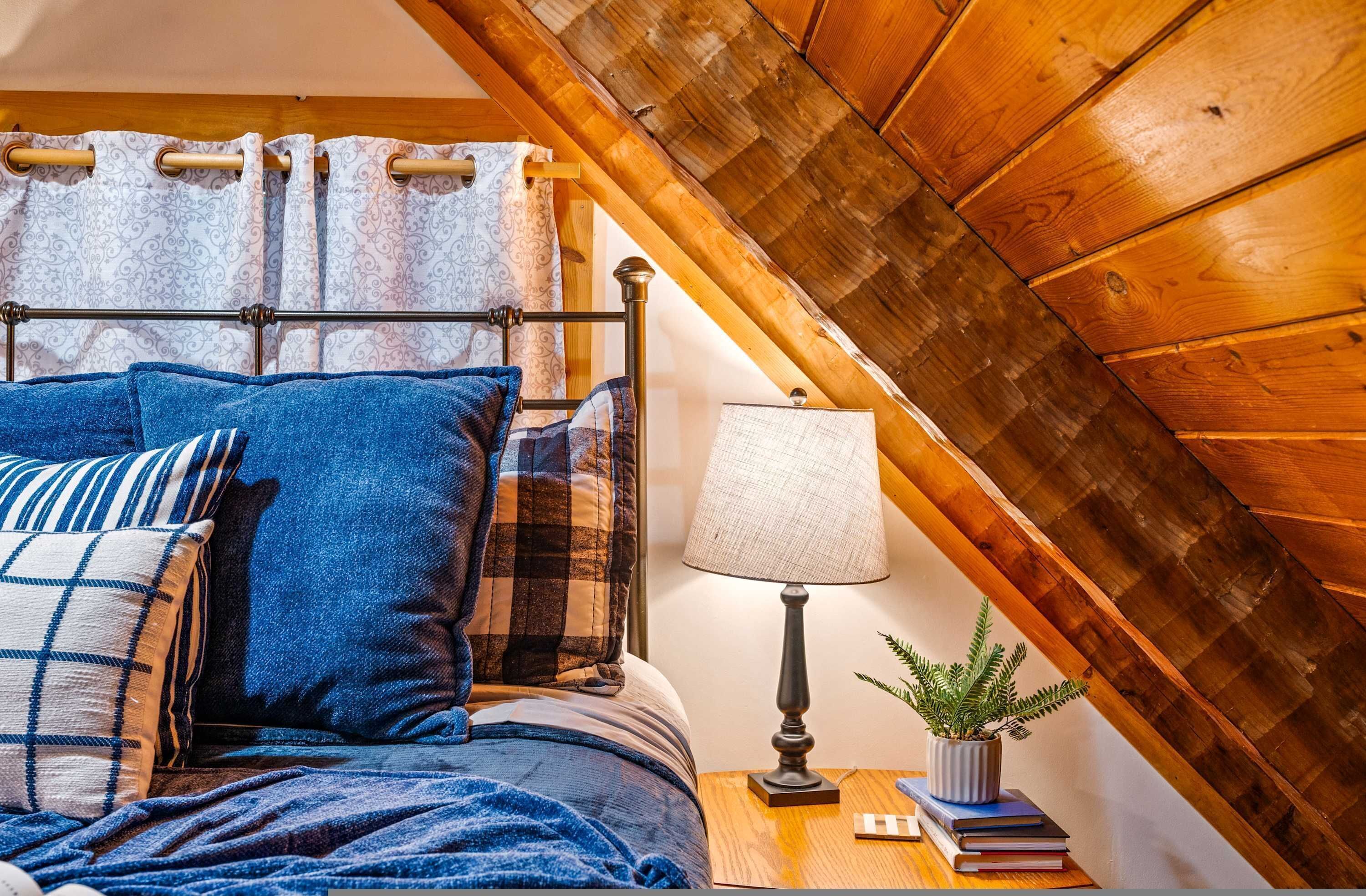 Ein Schlafzimmer mit Holzbalkendecke, einem Bett mit blauer Bettwäsche, einem Nachttisch mit Lampe und Pflanze sowie einem Vorhang mit Muster.