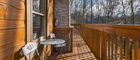 A wooden deck with a table and chairs, a stone wall, and a view of trees.