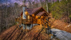 Une cabane dotée d’une cheminée en pierre, d’une terrasse en bois et d’un escalier menant à l’entrée.