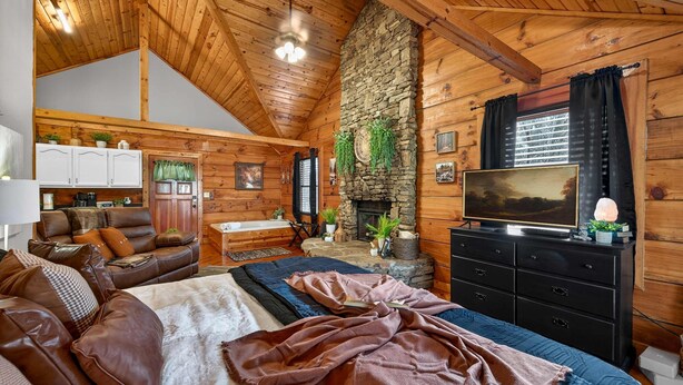 Interior de una cabaña de troncos con chimenea de piedra, techo de madera y una zona de estar acogedora con un sofá y televisor.