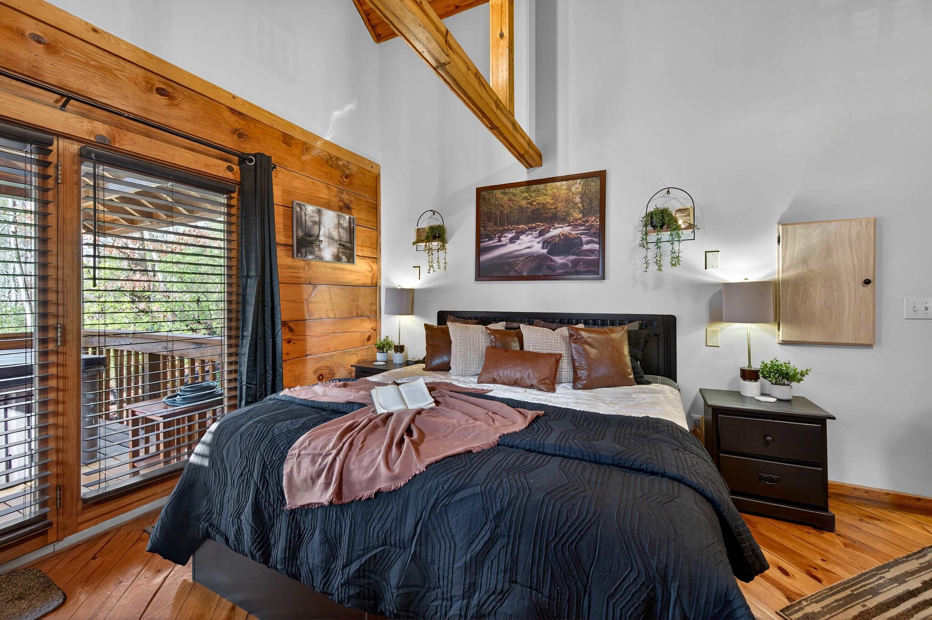 A bedroom with a large bed, wooden ceiling beams, a view through a window, and a nightstand with a lamp.