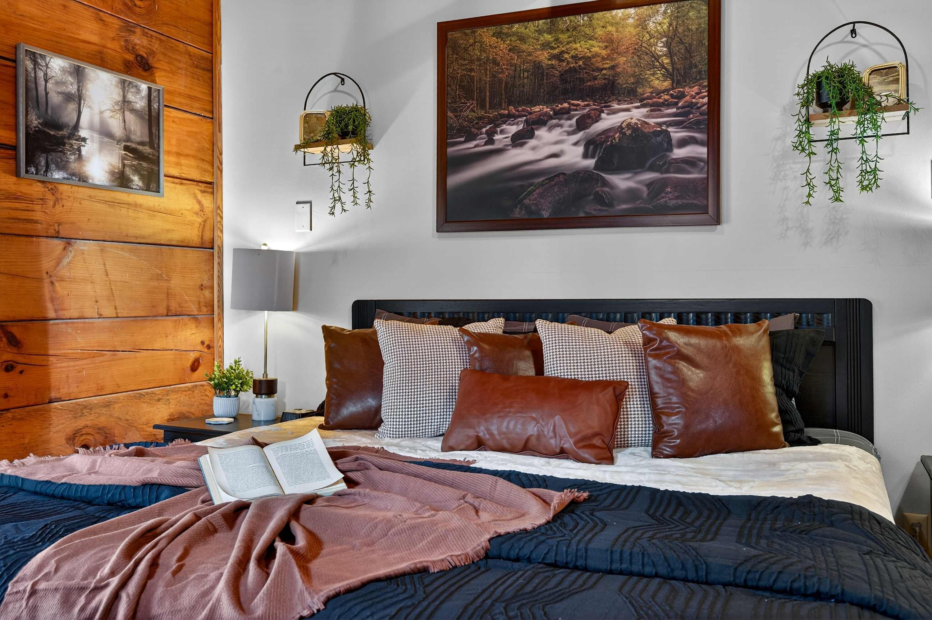 Een slaapkamer met een houten hoofdeinde, een groot bed met veel kussens, een nachtkastje met een lamp en twee aan de muur bevestigde planten.