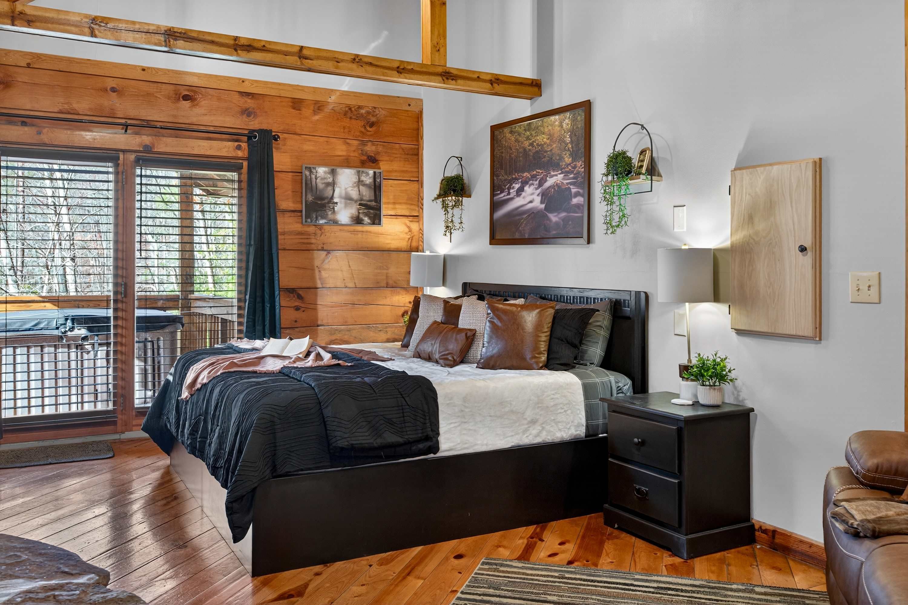 Un dormitorio con techo de madera, cama con estructura negra, mesita de noche y ventana con vistas a árboles.