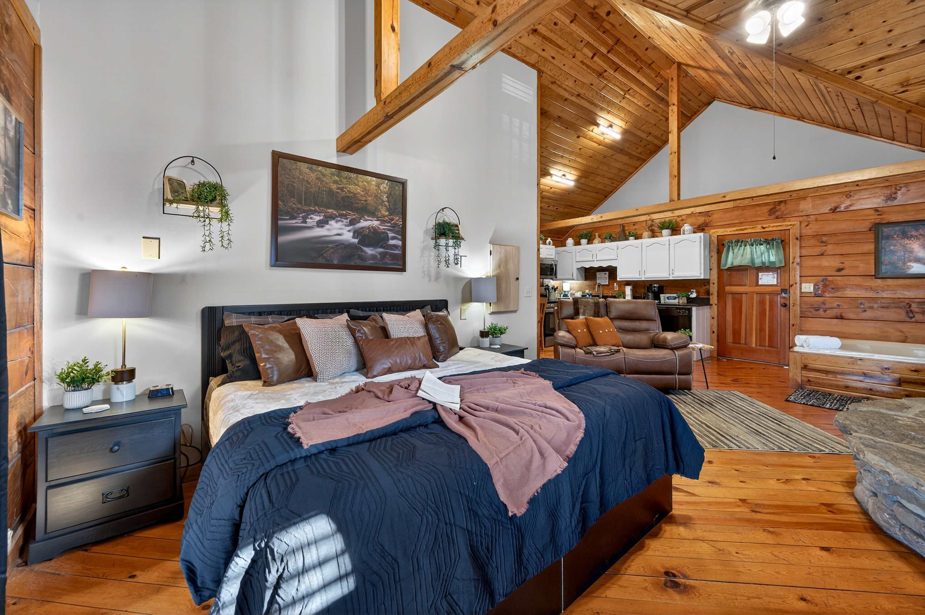 A bedroom with a large bed, wooden ceiling beams, a view into the kitchen, and a rustic cabin-style interior.