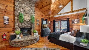 L’intérieur chaleureux d’un chalet avec une cheminée en pierre, des murs en bois, un lit, un téléviseur et un balcon offrant une vue imprenable.
