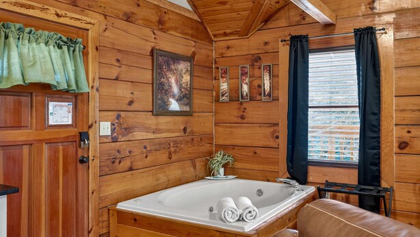 A wooden cabin bathroom with a large bathtub, a wooden door, a framed picture, and a window with curtains. - Hot Tub Location Cozy Studio Log Cabin Fireplace (Gatlinburg)