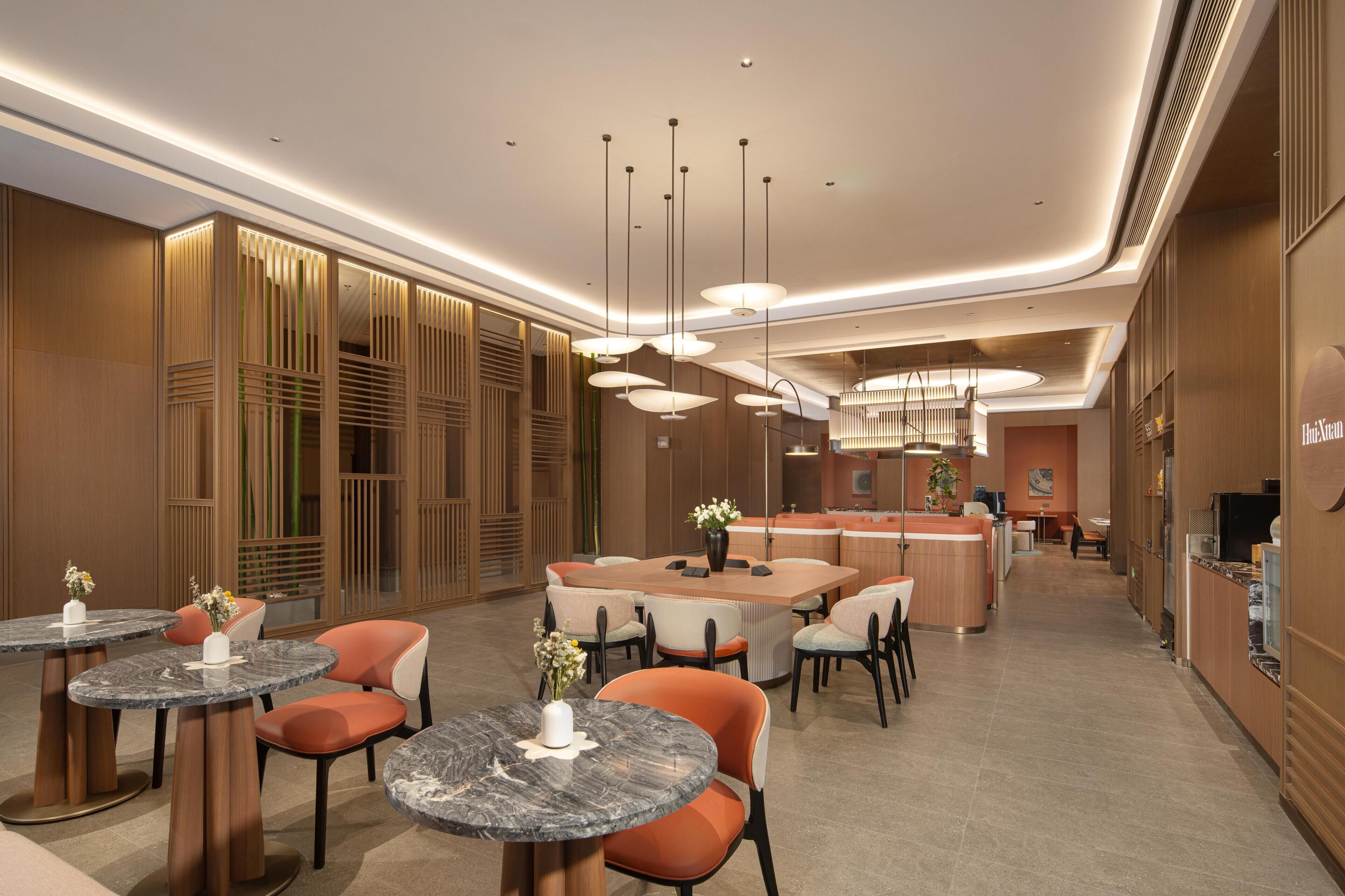A modern hotel lobby with round tables, orange chairs, a central island with seating, and a kitchen area in the background.