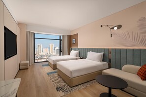 Ein Hotelzimmer mit zwei Betten, einem Schlafsofa, einem kleinen Tisch, einem Fernseher und Blick auf die Stadt.