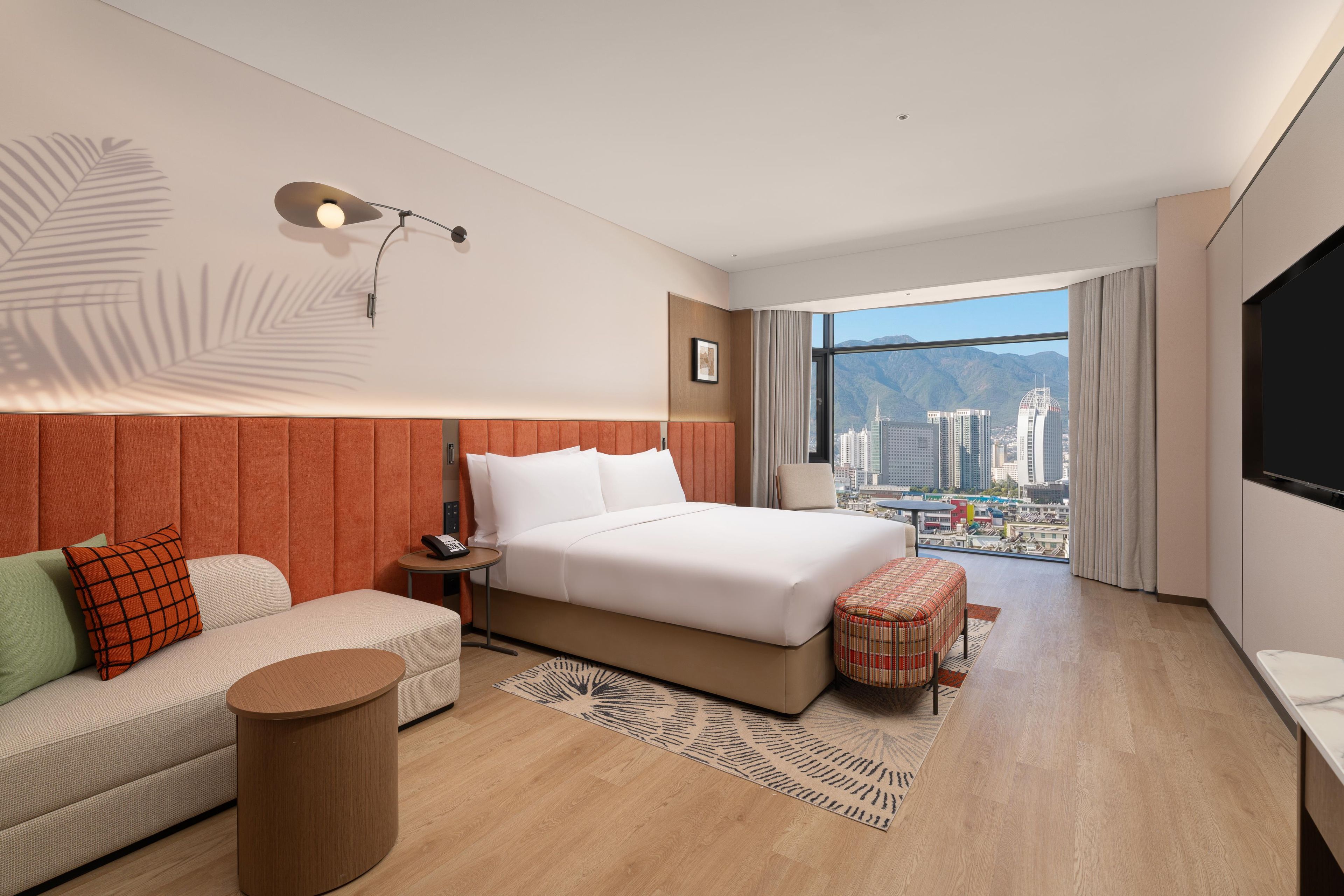 Habitación de hotel moderna con una cama grande, un sofá, una mesita, un televisor y vistas a la ciudad.