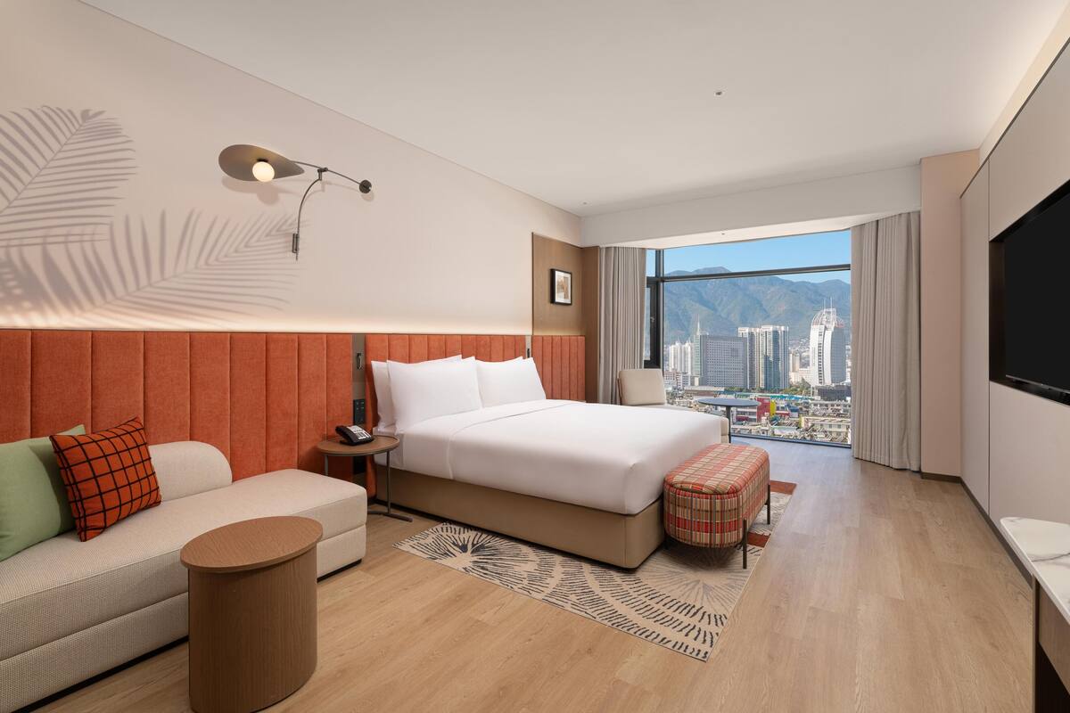 Une chambre d’hôtel moderne dotée d’un grand lit, d’un canapé, d’une petite table, d’une table d’appoint ronde, d’une télévision et offrant une vue sur le paysage urbain.