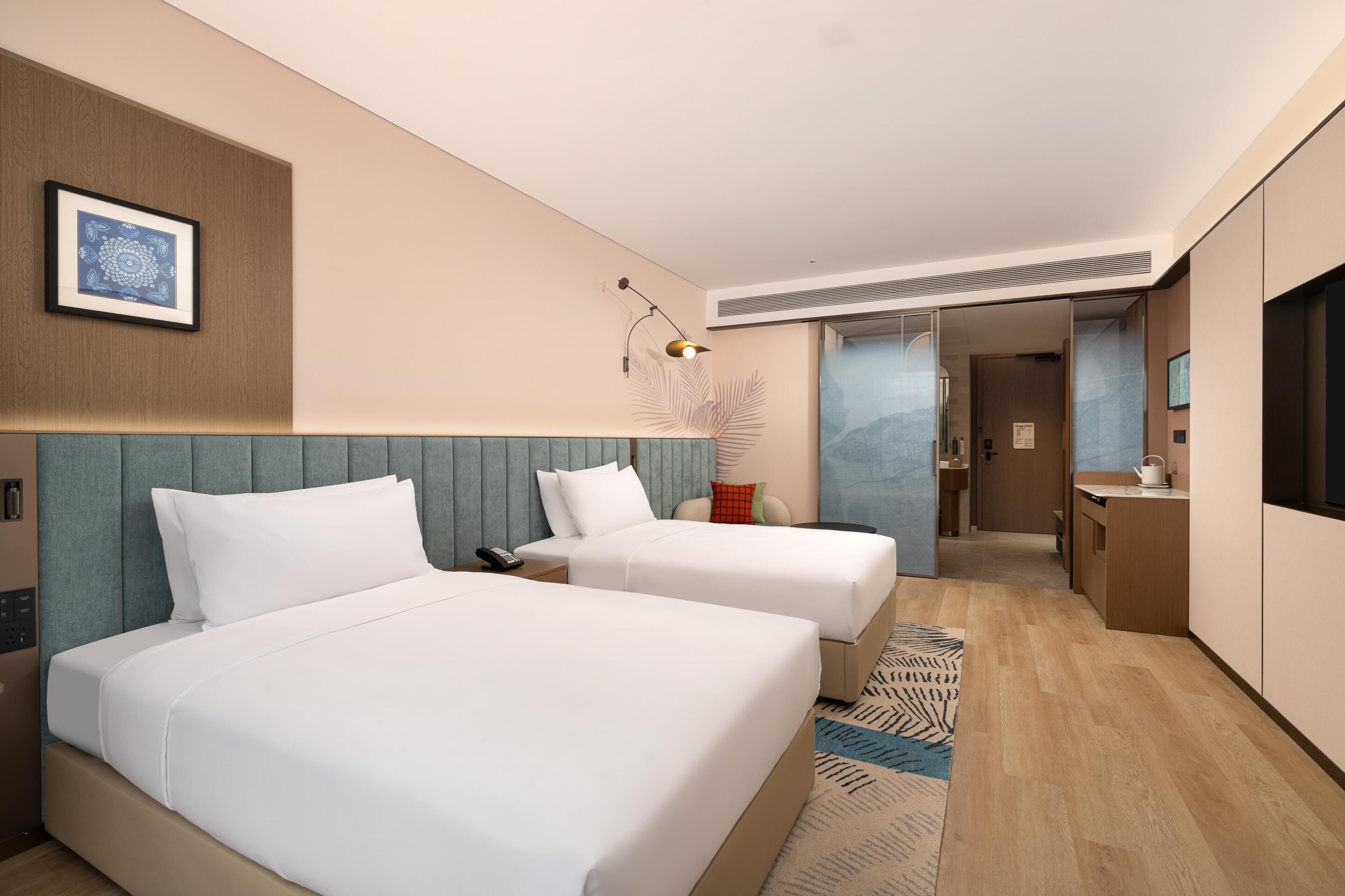 Habitación de hotel con dos camas, un escritorio, una silla, un televisor y un baño visible a través de una puerta abierta.