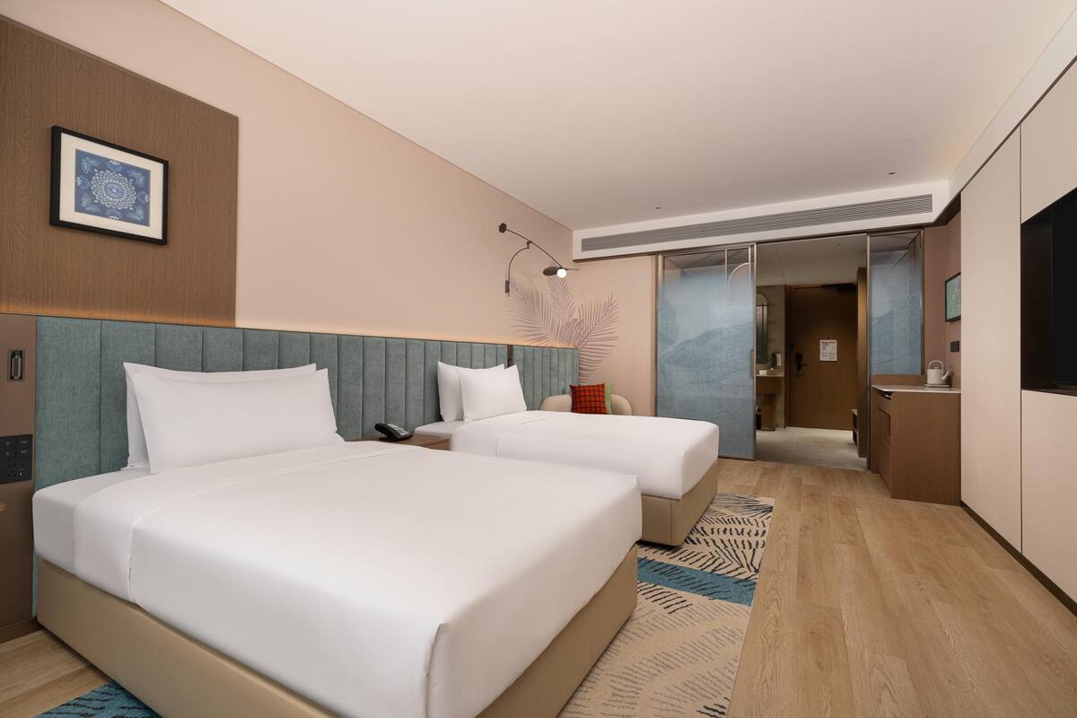 Une chambre d’hôtel avec deux lits, un bureau, une chaise, un miroir et une vue sur le couloir.