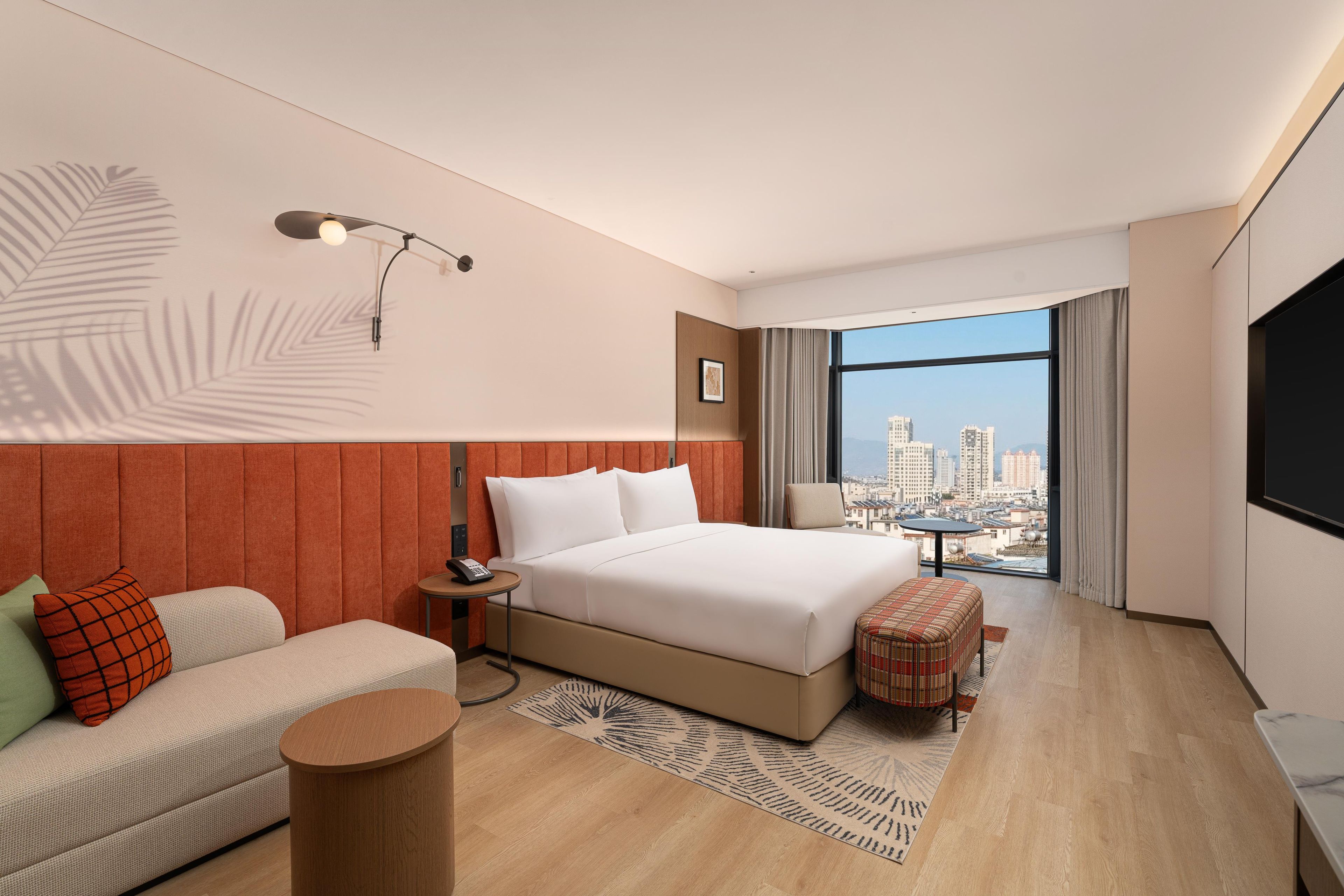 Ein modernes Hotelzimmer mit einem großen Bett, einer Sitzecke, einem kleinen Tisch, einem Schreibtisch und Blick auf die Stadt durch große Fenster.