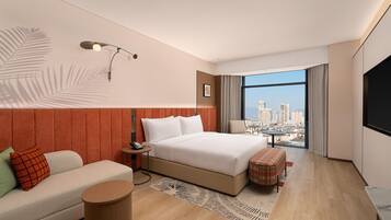 Ein modernes Hotelzimmer mit einem großen Bett, einer Sitzecke, einem kleinen Tisch, einem Schreibtisch und Blick auf die Stadt durch große Fenster.