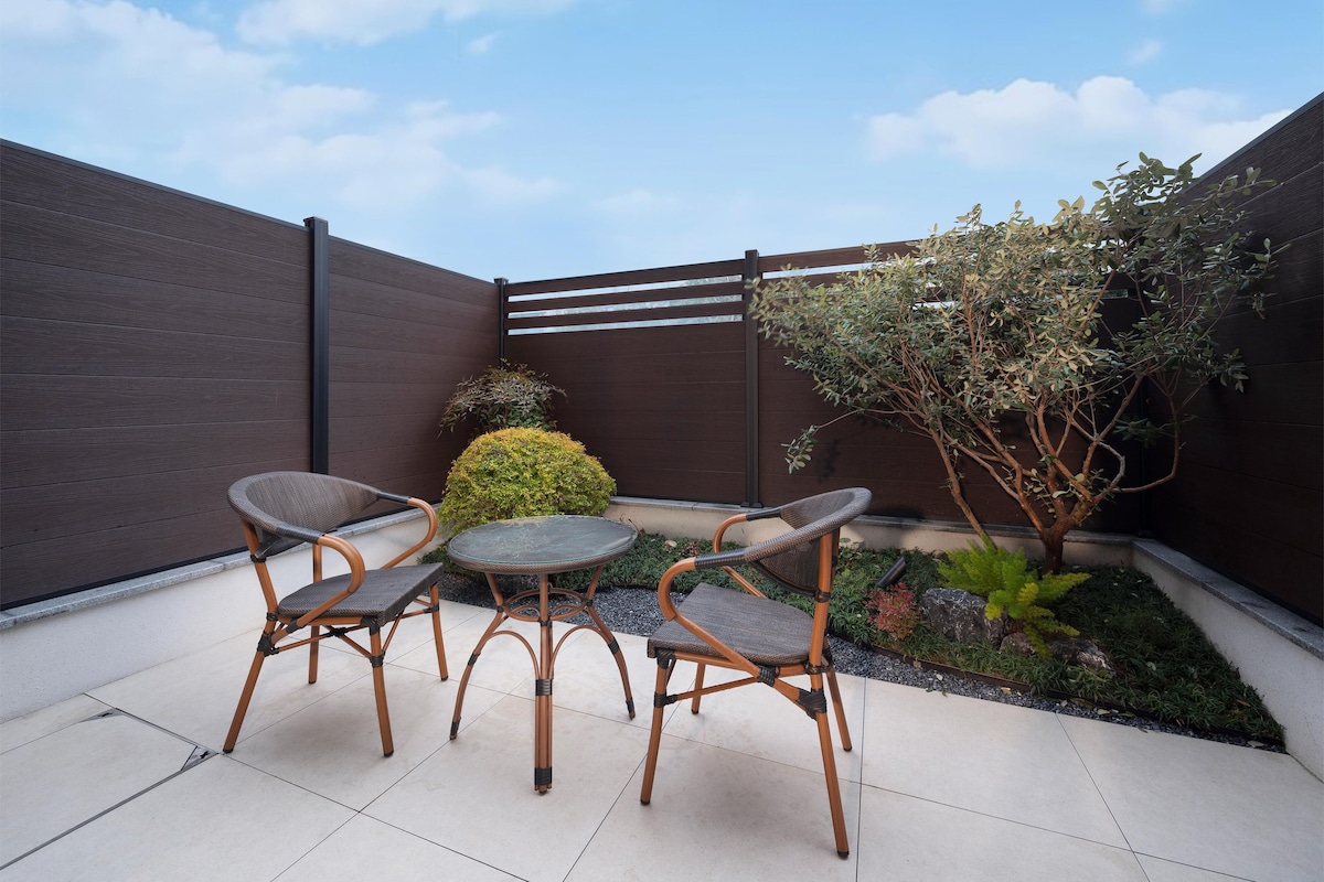 A patio area with a table and two chairs, a small bush, and a tree.