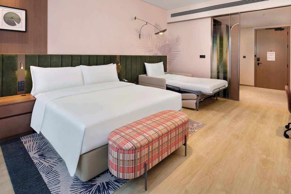 A hotel room with a large bed, a smaller bed, a bench, a desk, and a wall-mounted lamp.