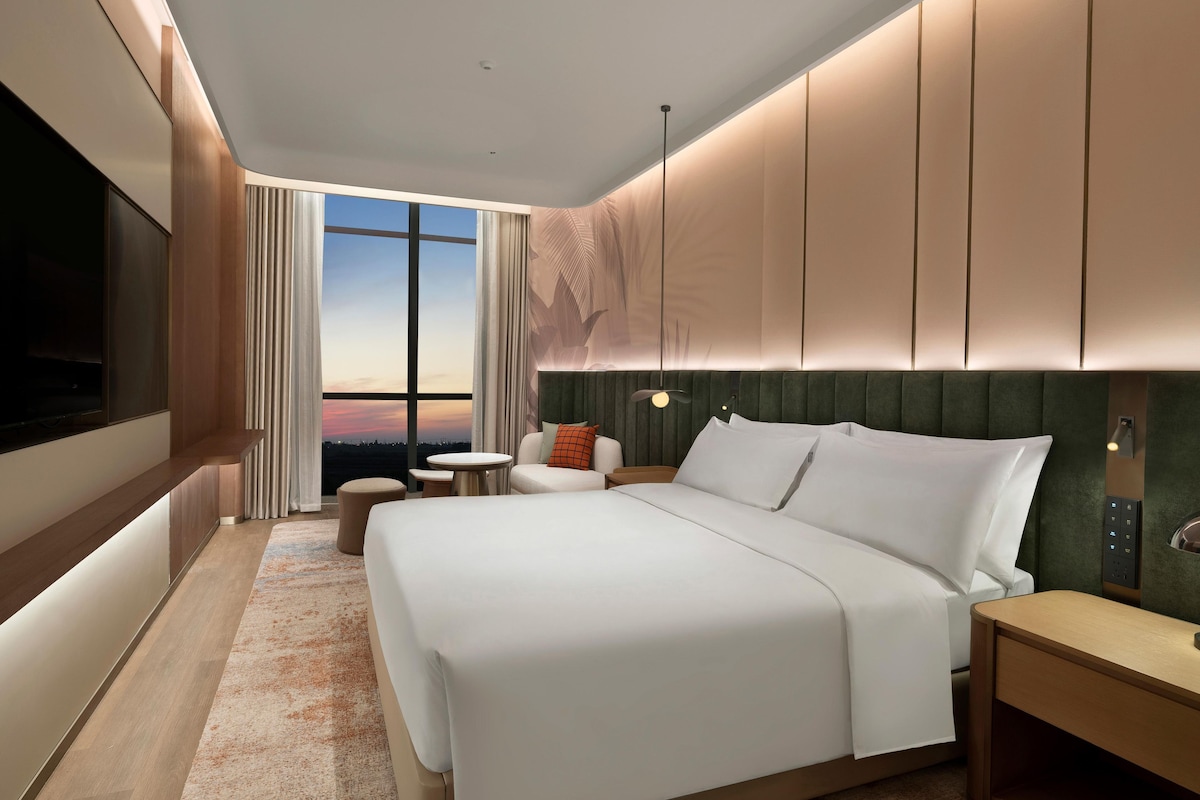 A modern hotel room with a large bed, a seating area with a table, and a large window offering a view.