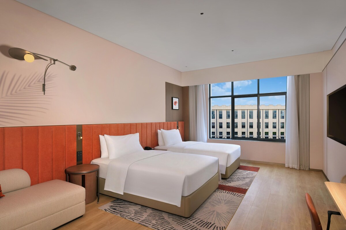 A hotel room with two beds, a sofa, a small table, a TV, and a window with a city view.
