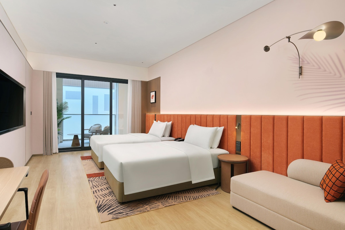 A hotel room with two beds, a sofa, a TV, a small table, and a balcony with a view.