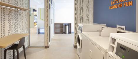 A laundry room with a wall sign that reads 'LET'S HAVE LOADS OF FUN,' a washing machine, a dryer, a trash can, and a wooden table with a chair.