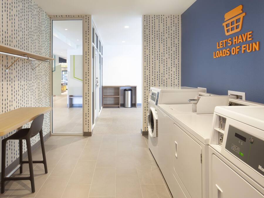 A laundry room with a wall sign that reads 'LET'S HAVE LOADS OF FUN,' a washing machine, a dryer, a trash can, and a wooden table with a chair.