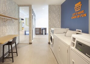 A laundry room with a wall sign that reads 'LET'S HAVE LOADS OF FUN,' a washing machine, a dryer, a trash can, and a wooden table with a chair. - Home2 Suites by Hilton Texarkana (Texarkana)