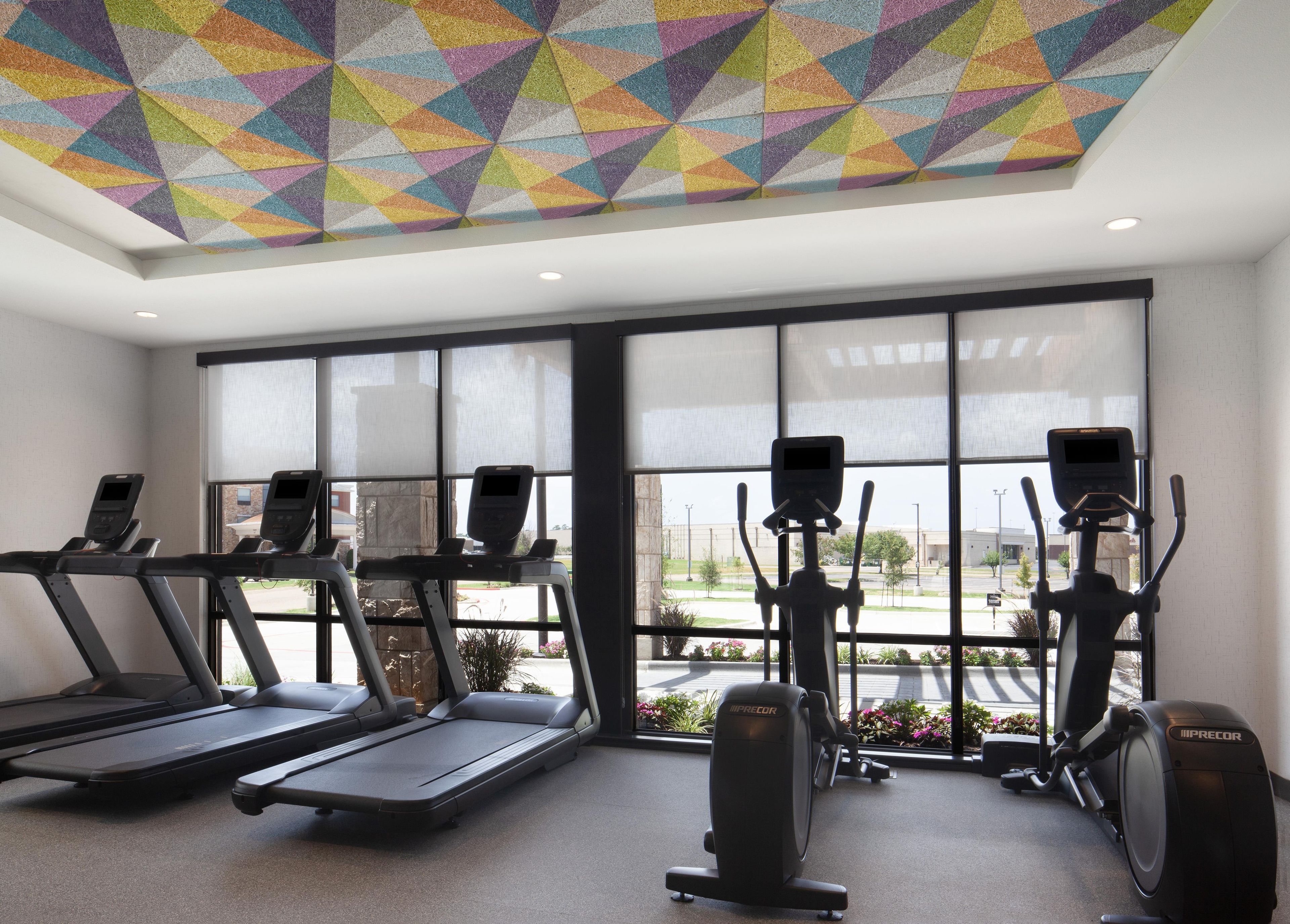 Ein Fitnessstudio mit Laufbändern, Crosstrainern und Ergometer, großen Fenstern und einer farbenfrohen, geometrischen Decke.