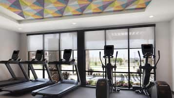 Ein Fitnessstudio mit Laufbändern, Crosstrainern und Ergometer, großen Fenstern und einer farbenfrohen, geometrischen Decke.