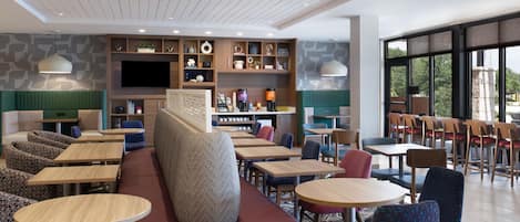 A modern hotel lobby with seating areas, a coffee station, and a large TV screen.