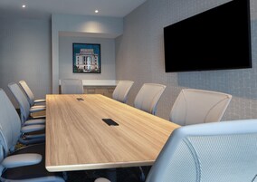 Un salón de conferencias con una larga mesa, seis sillas, un televisor montado y un cuadro en la pared.