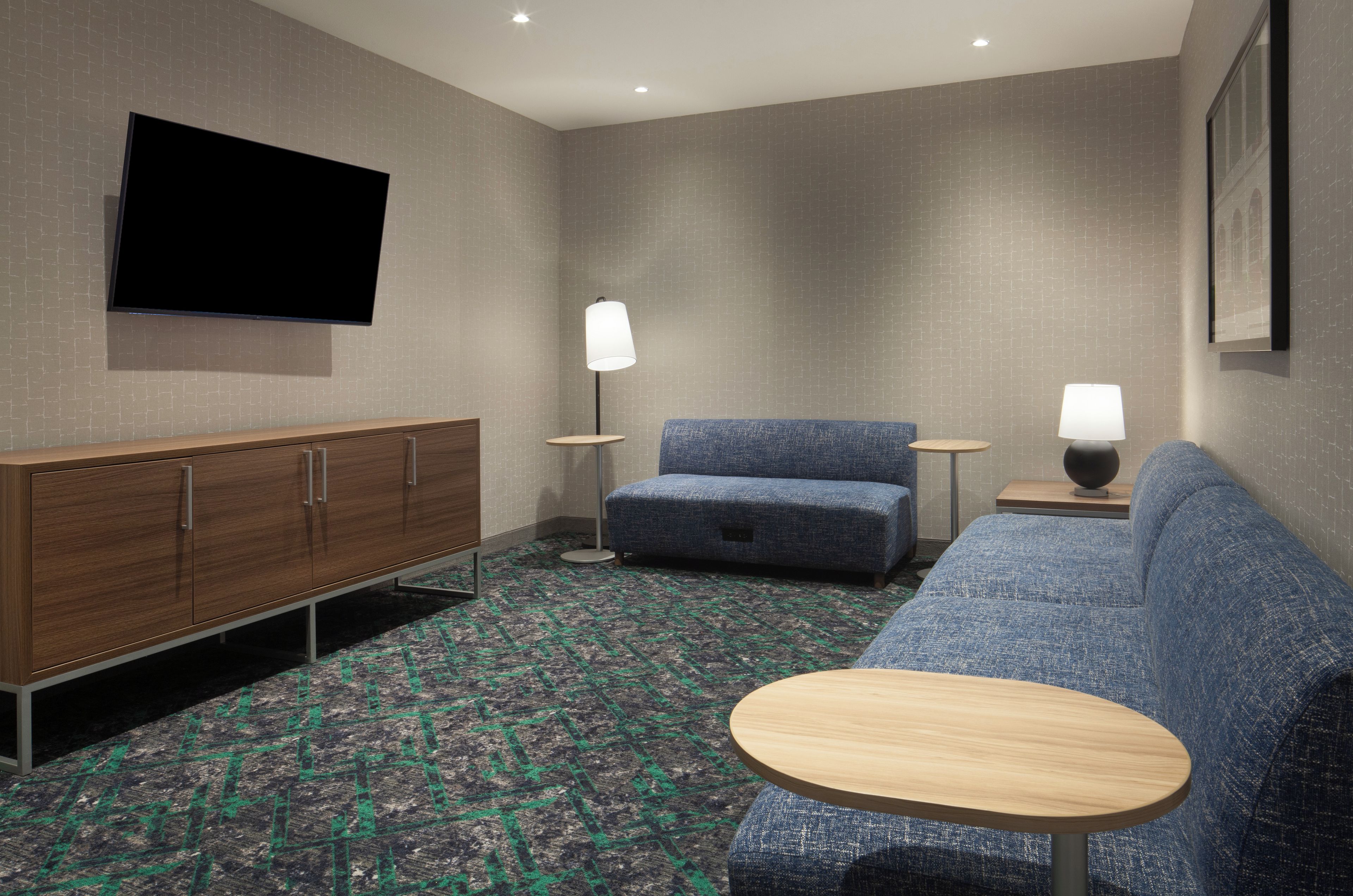 A hotel room with a flat-screen TV mounted on the wall, a wooden cabinet, a blue sofa, a round wooden table, and two floor lamps.