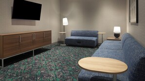 Ein Hotelzimmer mit einem an der Wand befestigten Flachbildfernseher, einem Holzschrank, einem blauen Sofa, einem runden Holztisch und zwei Stehlampen.