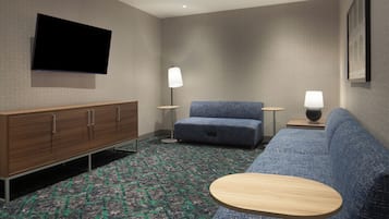A hotel room with a flat-screen TV mounted on the wall, a wooden cabinet, a blue sofa, a round wooden table, and two floor lamps.