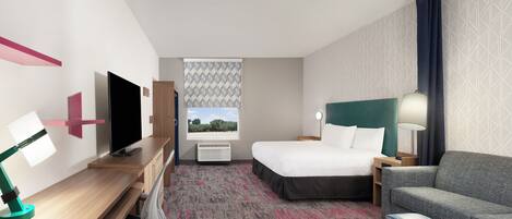 A hotel room with a bed, a desk with a TV, a chair, a sofa, and a patterned carpet.