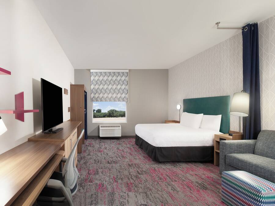 A hotel room with a bed, a desk with a TV, a chair, a sofa, and a patterned carpet.