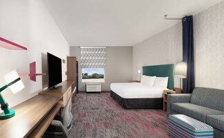 Habitación de hotel con cama, escritorio con televisor, una silla, un sofá y alfombra estampada.
