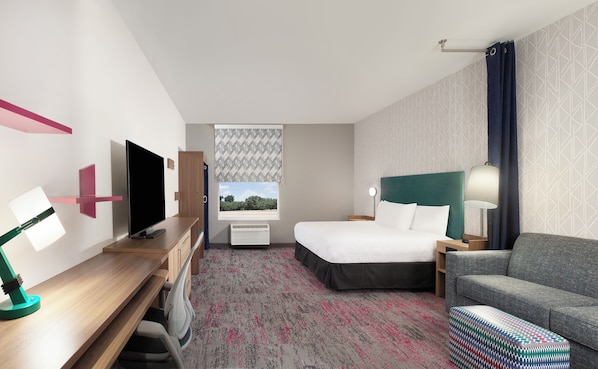 A hotel room with a bed, a desk with a TV, a chair, a sofa, and a patterned carpet.